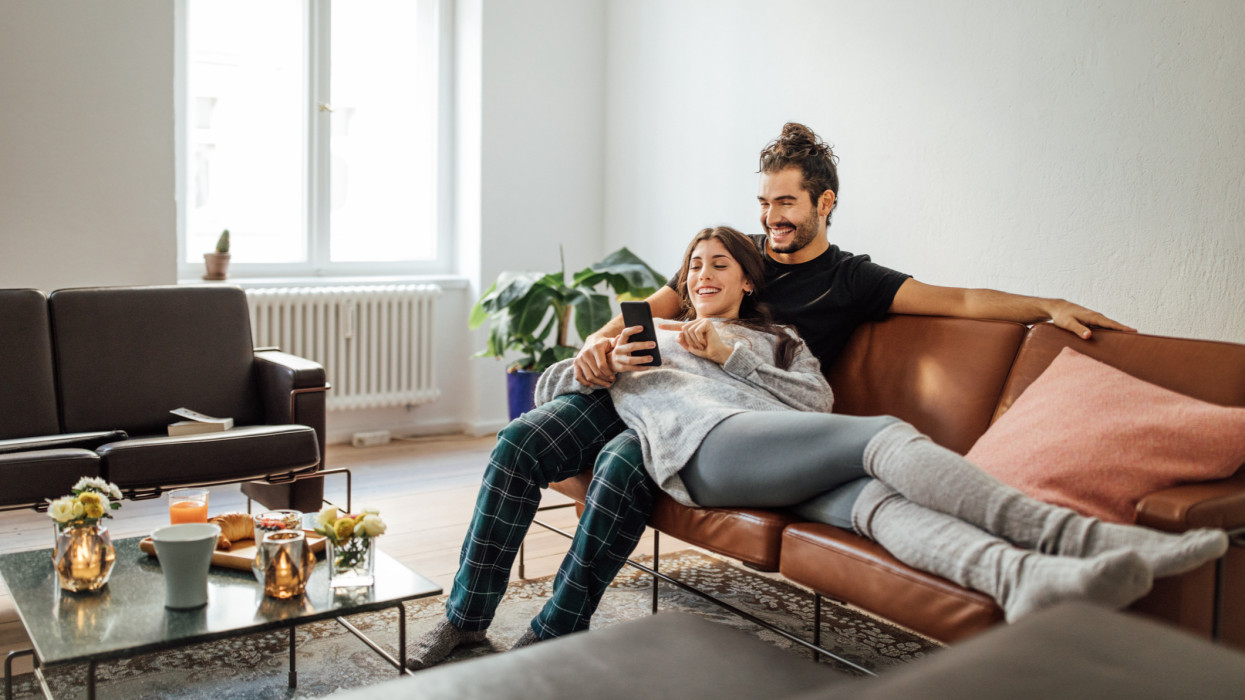 Smiling young woman using mobile phone while reclining on boyfriend sitting on sofa at home