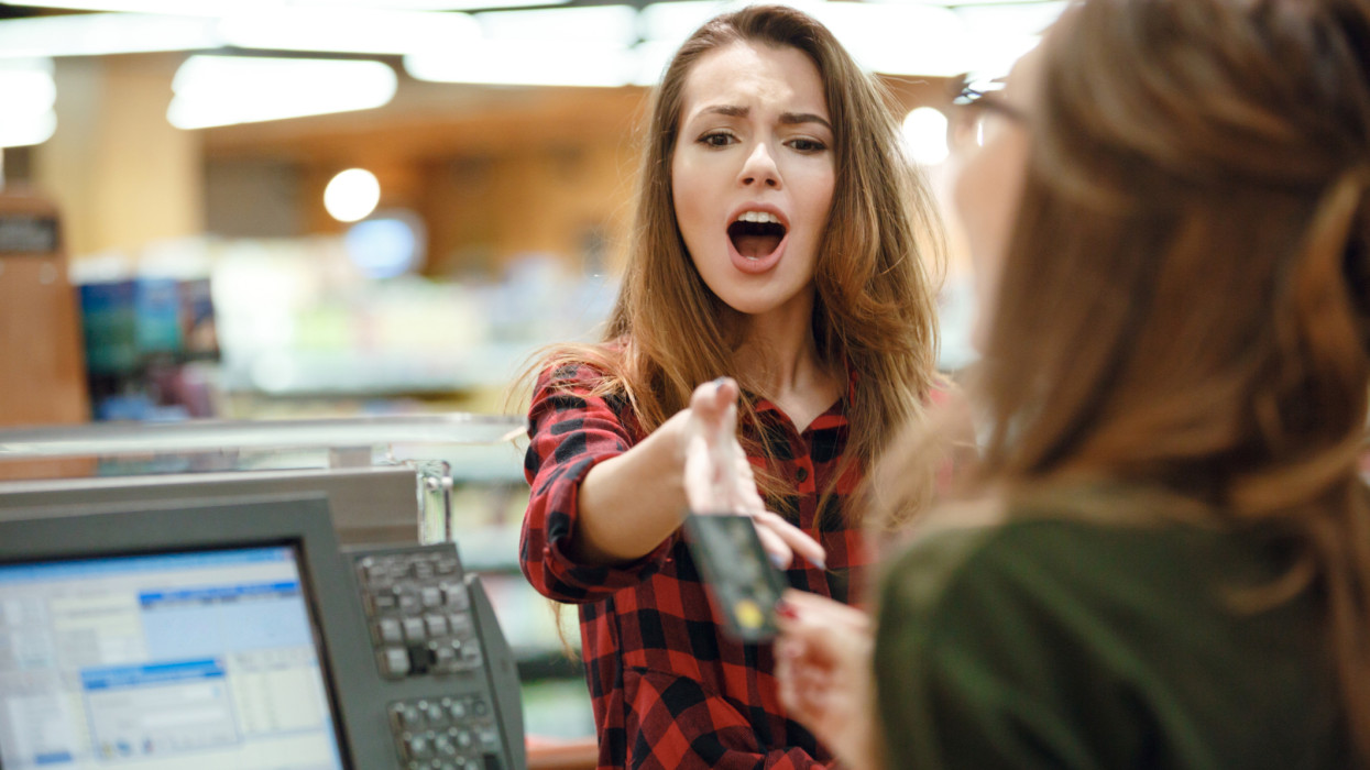 Image of confused young lady standing in supermarket shop near cashiers desk looking at credit card.